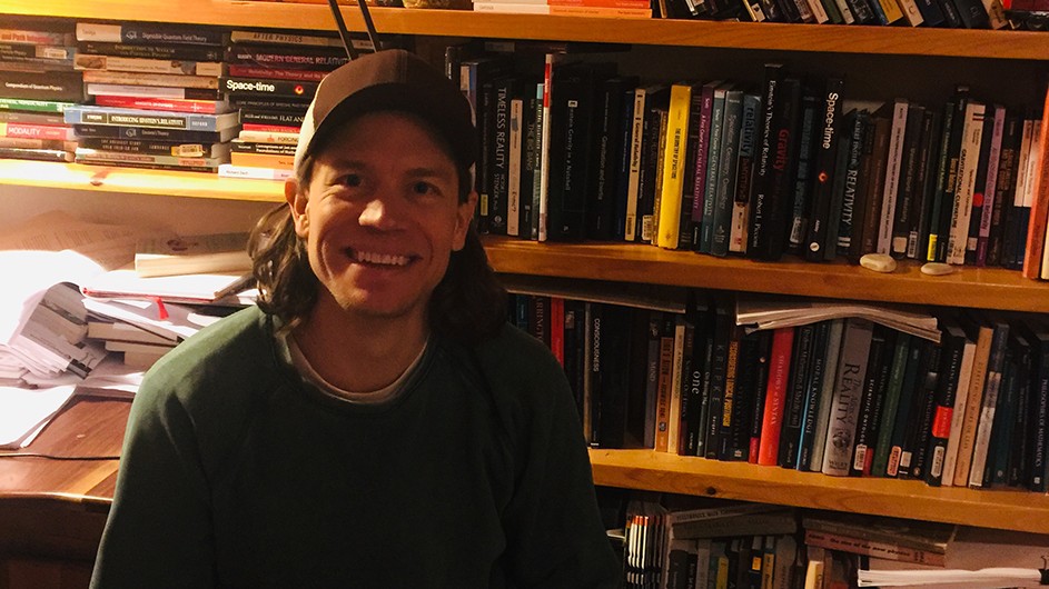 Philosophy Professor Justin Clarke-Doane has been reading physics textbooks lately. Justin Clarke-Doane: a man wearing a baseball cap sits in front of a desk with papers and full bookshelves.