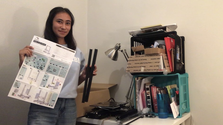 A girl with dark hair holds an instruction sheet and stands next to a desk containing her engineering kit.