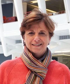Lynne Sagalyn wears a watermelon colored sweater and a multicolored neck scarf while smiling directly into camera lens