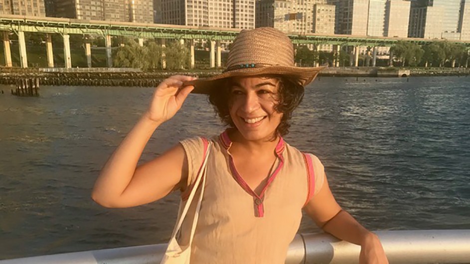 Mana Kia: a smiling woman wears a hat and stands in front of water and buildings.