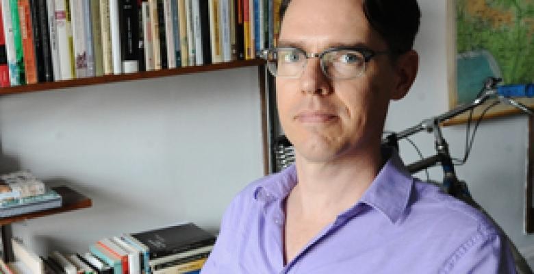 Gregory Mann looks into camera lens wearing eyeglasses and a lavendar collared shirt 