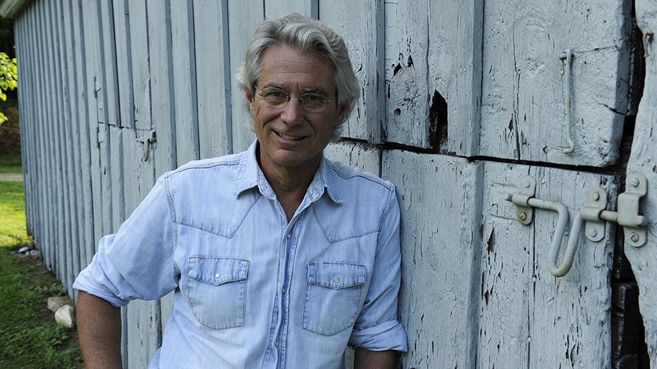 A man with white hair and eyeglasses, wearing a jean shirt and leaning against a white wooden wall.