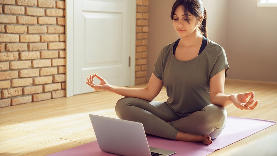 young woman with brown hair in pony tail in yoga pose, sitting cross-legged with arms out to sides and fingers touching. She is looking at a laptop while sitting on the floor..
