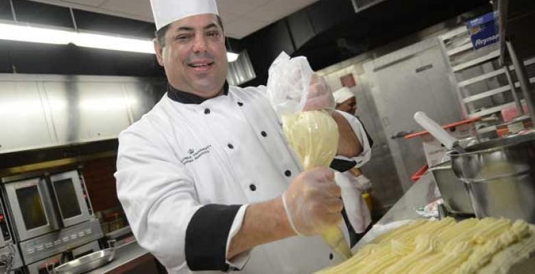 Standing in an industrial styled kitchen Michael Demartino dressed in wearing a white chef's hat and jacket while squeezing cookie dough from pressing bag onto sheet