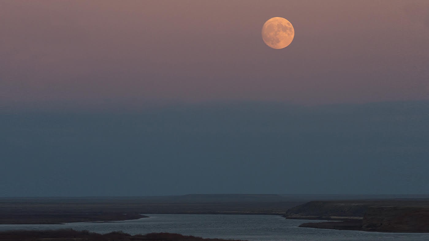 A full pinkish colored moon against a blue and pink sky with barren land and water inlet below.