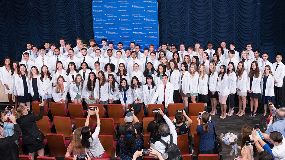 Dozens of dental students in white coats smile and pose in front of blue Columbia signage as their friends and family take photos of them at Columbia University Irving Medical Center.