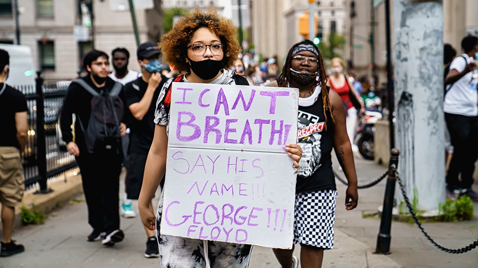 A woman wearing a face covering, holding up a sign--which reads, "I can't breathe! Say his name. George Floyd!!!--walking with a group of protestors..