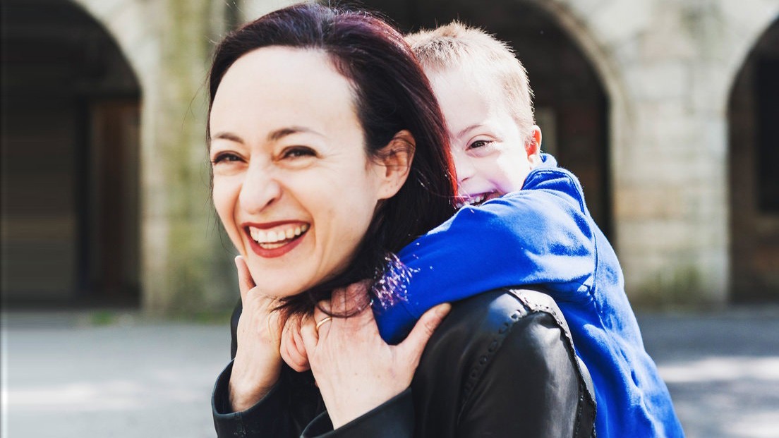 Professor Rachel Adams and her son, Henry. A smiling woman holding a child on her back, who is wearing a blue top.