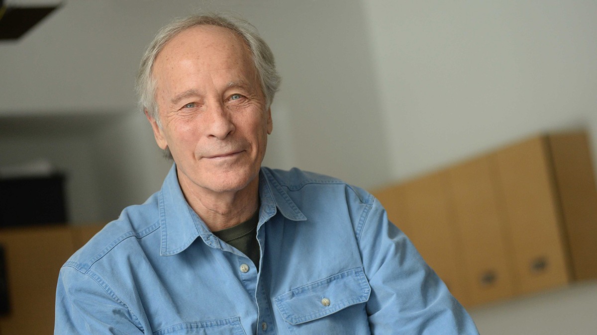 Richard Ford: A man with white hair, wearing a light blue denim shirt, smiles into the camera. 