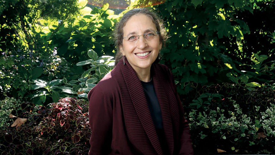 A smiling woman with glasses wears a maroon sweater and sits before a background of sunlit greenery.