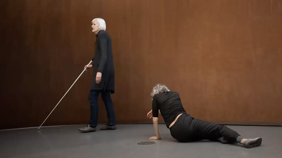 A woman with white hair and a cane walks behind a person leaning on the ground.