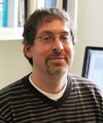 Jeffrey Shaman faces camera wearing black and gray striped vneck sweater with a white t-shirt lining the top color. Sharman is wearing black wire rimmed eyeglasses.