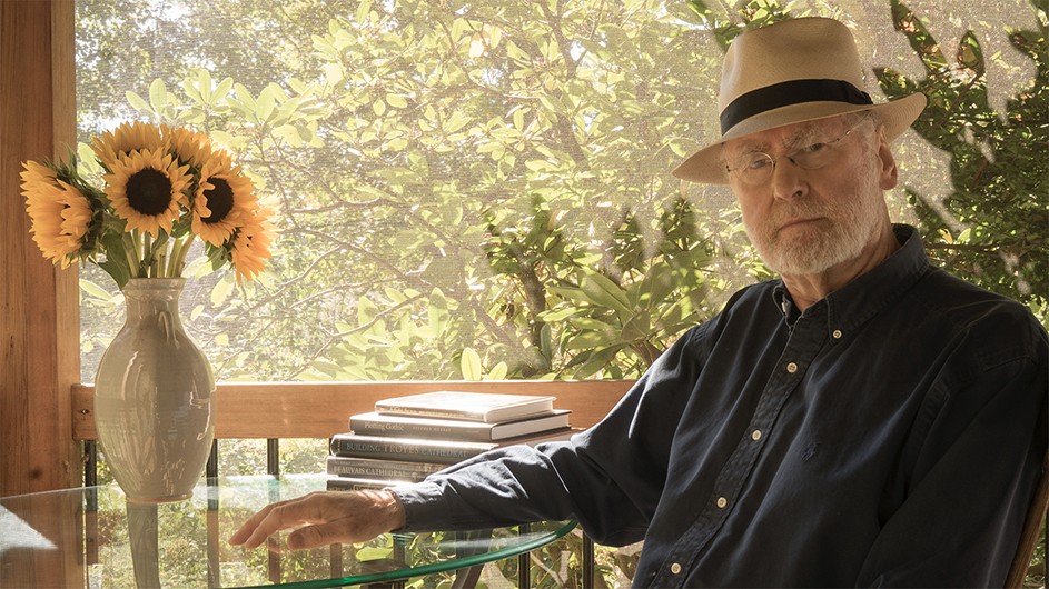 A man wearing a hat sits at a table with a vase of sunflowers.