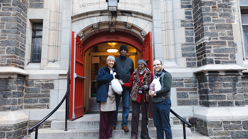 Four people holding Thanksgiving turkey donations and standing on the steps of Broadway Community Church in front of an open red door in Morningside Heights, New York City.
