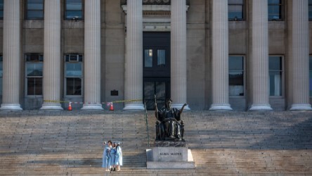 Three students in caps and gowns posing in front of a statue on the steps with a columned building in the background.