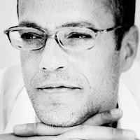black and white headshot of a man in glasses with his chin resting on his hands