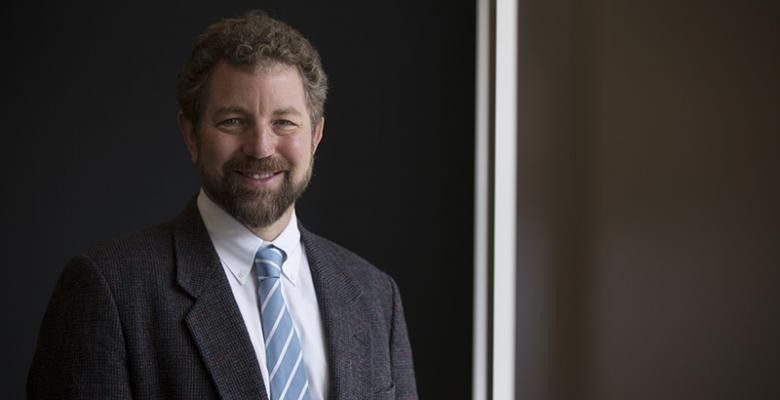 Tommy Vaughan dressed in a dark blazier with a white button down and light blue tie.