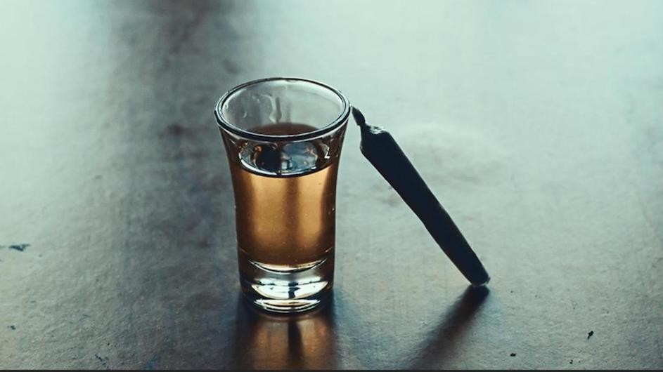 A shotglass of alcohol next to a marijuana cigarette.