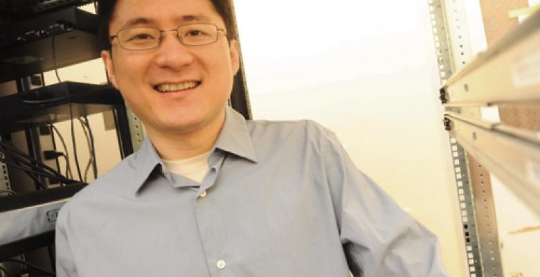 Prof Junfeng Yang smiles into camera wearing dark wired rimmed eyeglasses and a gray buttoned down shirt with a white s shirt peeping out at the neck