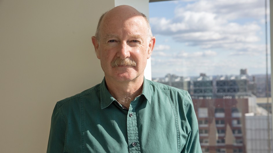 Man with grey hair and mustache in green shirt standing at window with NYC skyline
