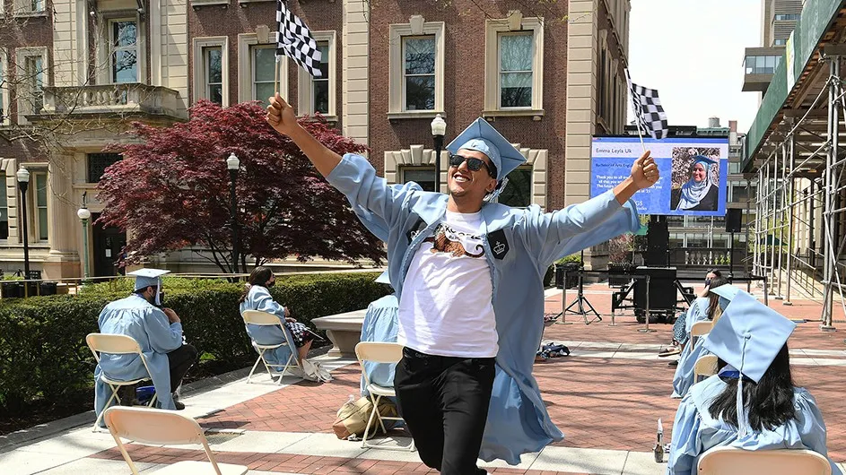 A student in Columbia graduation gown strides happily at a virtual commencement ceremony. 