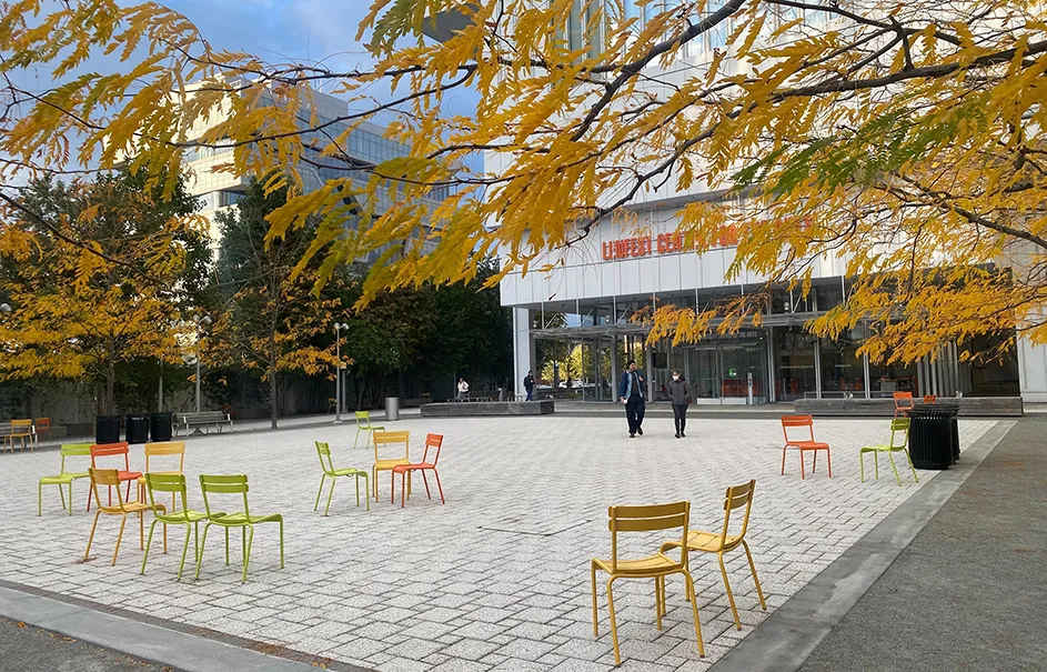 A dozen yellow, orange, and green chairs casually set up on the small square in front of the Lenfest Center for the Arts with yellow leaves prominently in view from the nearby trees at Columbia Manhattanville on a sunny autumn day.