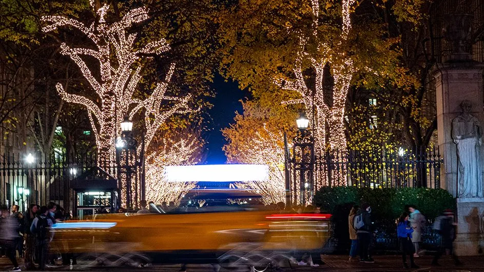 A yellow cab speeds in front of College Walk with trees lit for Christmas.