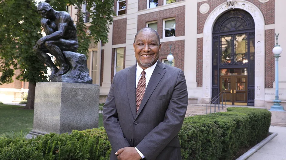 A man in a suit and tie, standing against the backdrop of a university building and a statue of a man sitting and thinking.