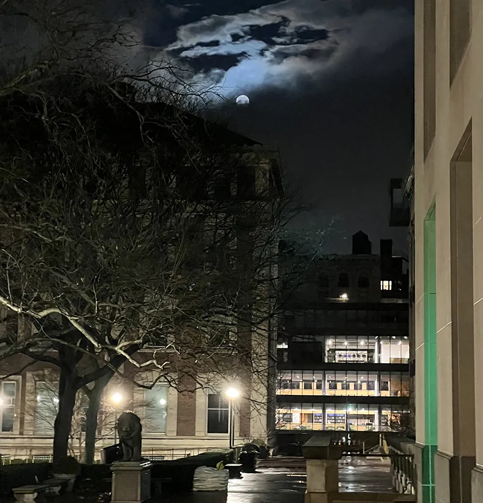 The bright full moon glows above Columbia University's Havemeyer and Math Plaza near Scholars' Lion statue with a view of Barnard College's illuminated library in the distance.