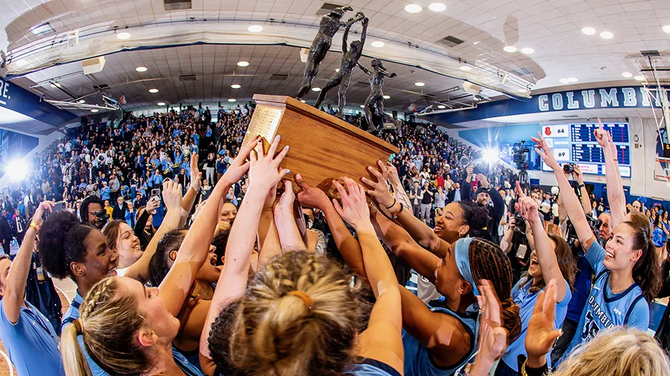 Several Columbia women's basketball players smile and hold up the Ivy League championship trophy in front of hundreds of fans in the bleachers of Levien Gym at Columbia University.