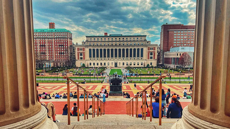 A view flanked by two Low Library center columns toward Alma Mater statue and Butler Library with dozens of students sitting on Low Library steps and South Lawn fields on a cloudy day at Columbia University.