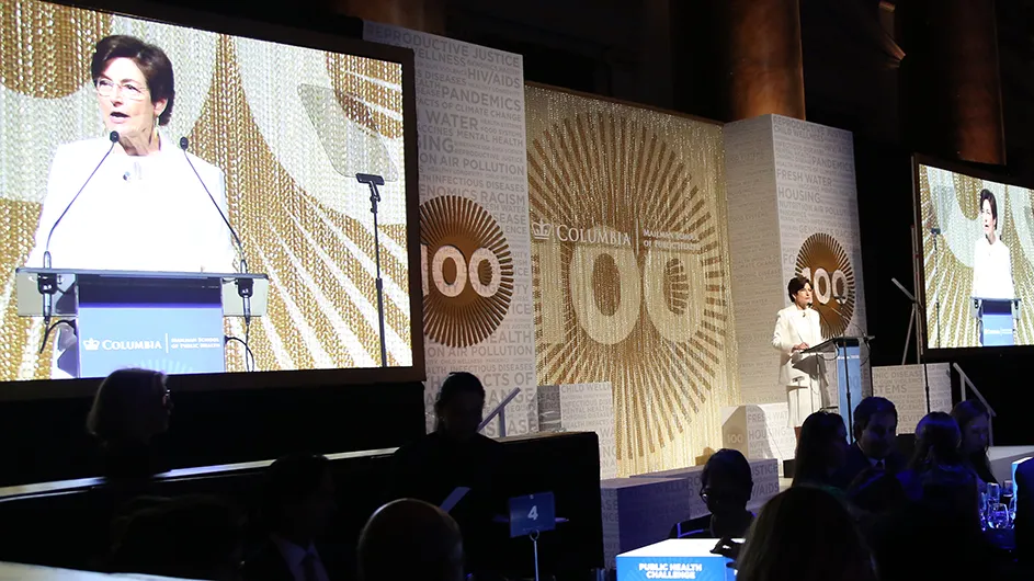 Columbia Mailman Dean Linda P. Fried speaking at a podium on stage with Columbia Mailman centennial signage flanked by two big screens of the dean speaking at the Columbia Mailman Centennial Gala.