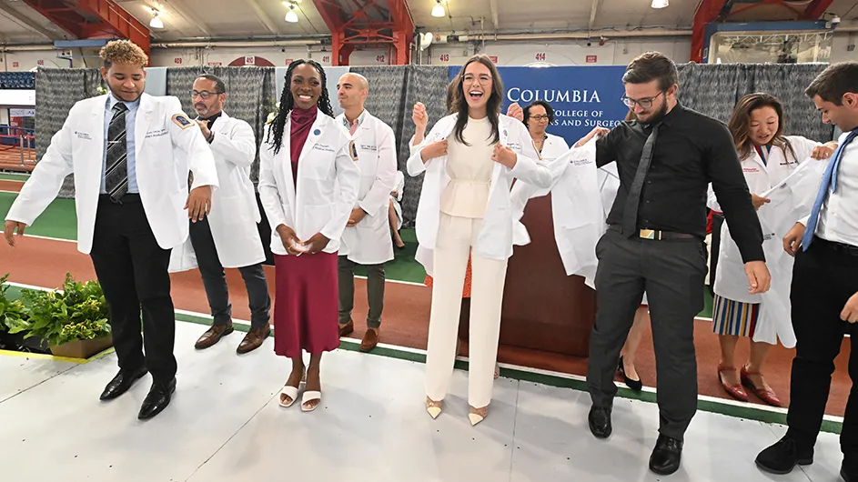 Five Columbia medical students are putting on their white medical coats in front of six faculty members in white coats at Columbia's Vagelos College of Physicians and Surgeons' White Coat Ceremony.