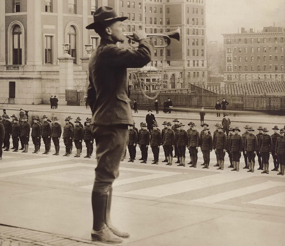 A student in 1918 wearing a WWI military uniform playing the bugle in front of dozens of uniformed students standing at attention on Low Plaza at Columbia University.  