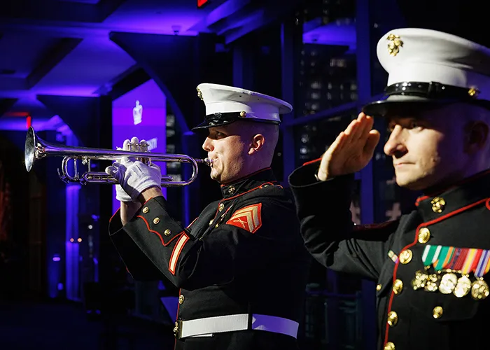 A trumpeter in dress blues plays as another service member with a chest full of honors salutes.