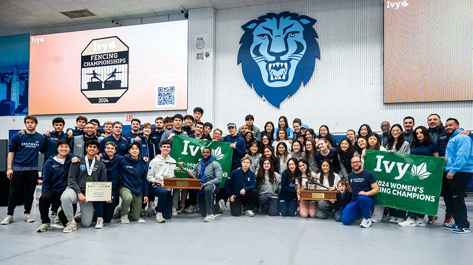 A group of Columbia fencers celebrate while posing and holding two Ivy League championship trophies and signage at Columbia University.