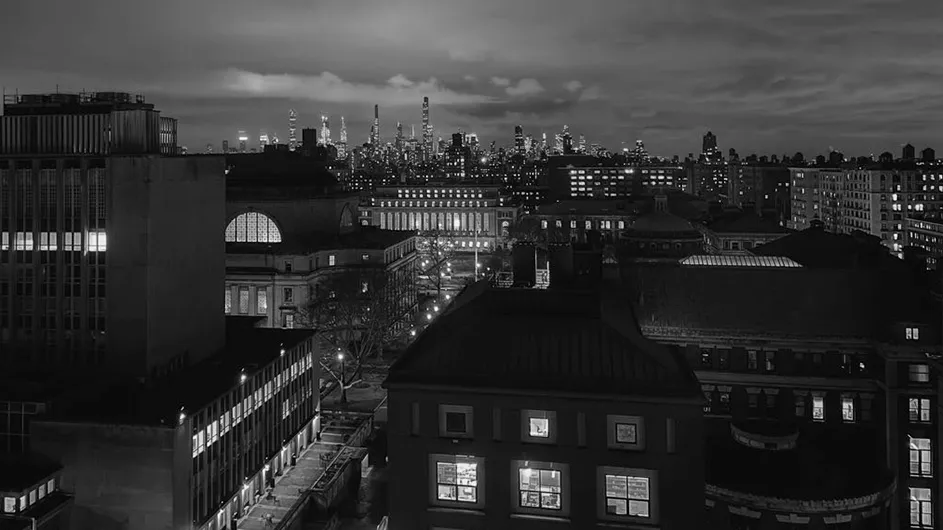 Columbia University's illuminated Morningside campus with New York City's skyline in the distance on a cloudy night.