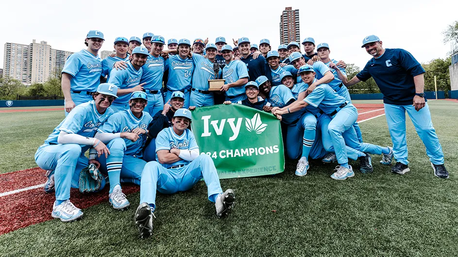 Several men's baseball players in Columbia blue uniforms smile and pose with a trophy and Ivy League 2024 Champions sign at Columbia's Baker Athletics Complex.