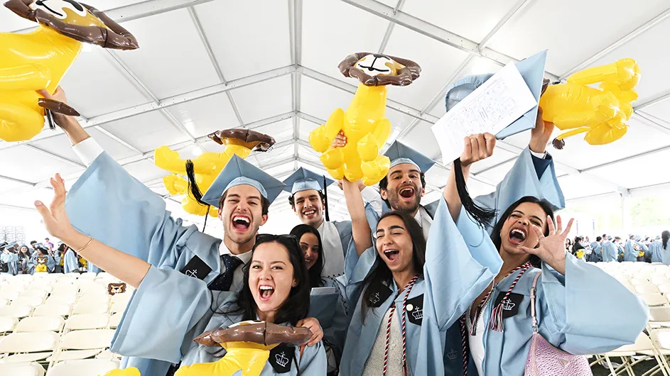 Seven students wearing light blue graduation regalia cheer and roar while holding inflatable balloon lions at Columbia College Class Day.