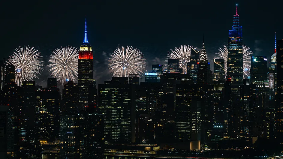 New York City skyline of Empire State building, Chrysler Building, and other skyscrapers with white fireworks illuminating the sky on July 4th. 