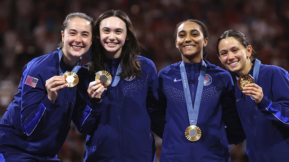 Four fencers in blue USA uniforms smile while holding their Olympic gold medals on the podium in a packed stadium in Paris.