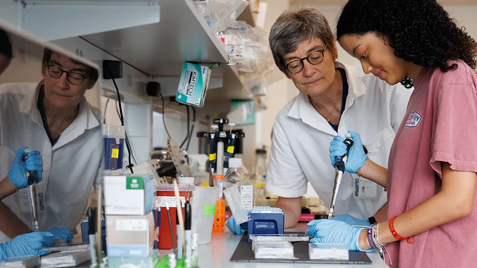 A student wearing blue lab gloves uses a scientific instrument to put a specimen onto a lab slide for observation as a professor looks on at Columbia's Zuckerman Institute. 