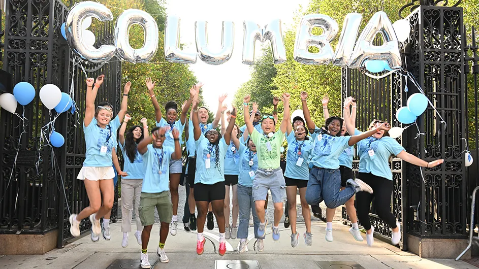 Several students in light blue shirts jump and cheer at the entrance of Columbia University, decorated with balloons displaying Columbia's name.