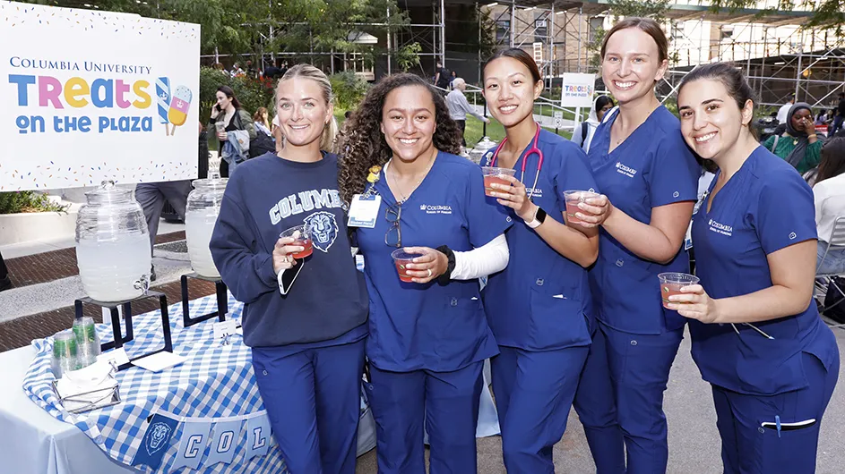 Five students and staff in Columbia blue scrubs smile and hold up glasses of lemonade at Treats on the Plaza at Columbia University Irving Medical Center's Haven Plaza near a table with lemonade. 