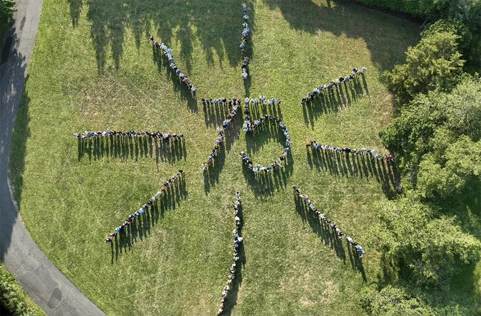 Dozens of people stand in a number 75 formation with eight rays of sun extended from the center at Columbia University Lamont-Doherty Earth Observatory in Palisades, New York, on a sunny day.  
