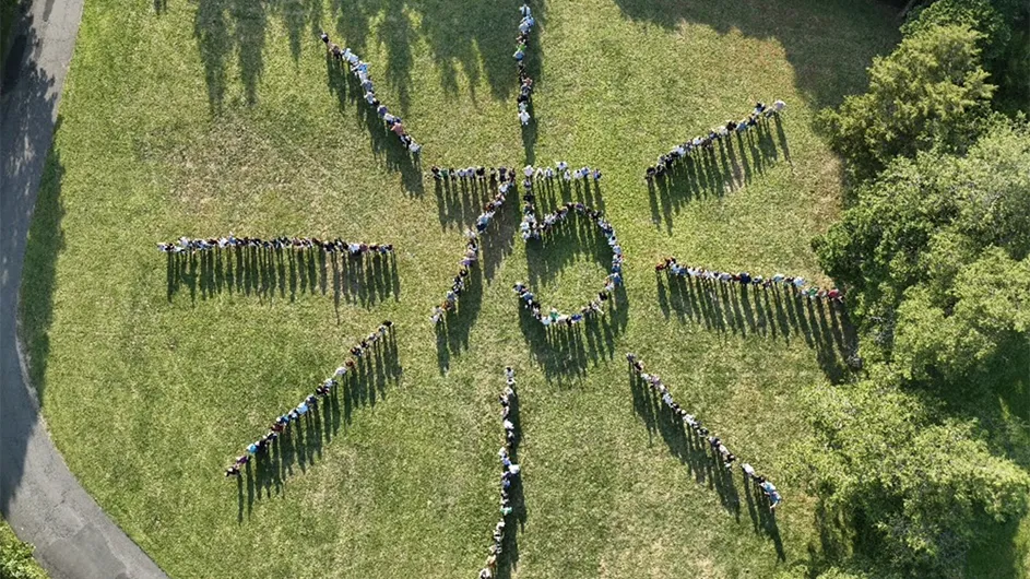 Dozens of people stand in a number 75 formation with eight rays of sun extended from the center at Columbia University Lamont-Doherty Earth Observatory in Palisades, New York, on a sunny day.  