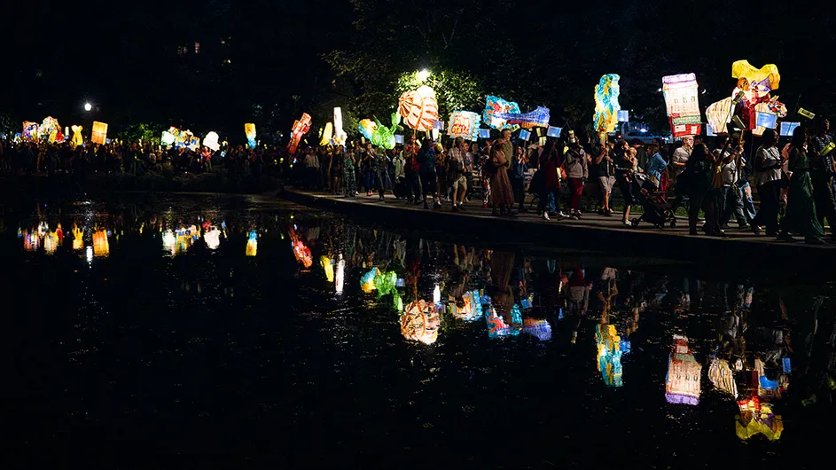 Dozens of people on a parade through Morningside Park carry colorful illuminated handmade lanterns which are reflected in the pond on a warm night in New York City. 