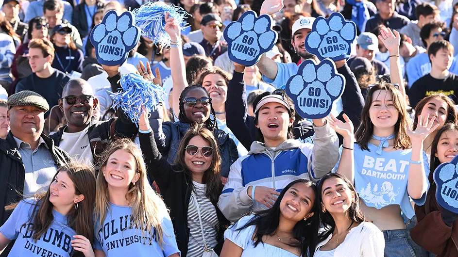 Several college students and alumni dressed in light blue Columbia shirts and holding up "Go Lions!" paw signs cheer while posing for the camera at Columbia University Homecoming. 