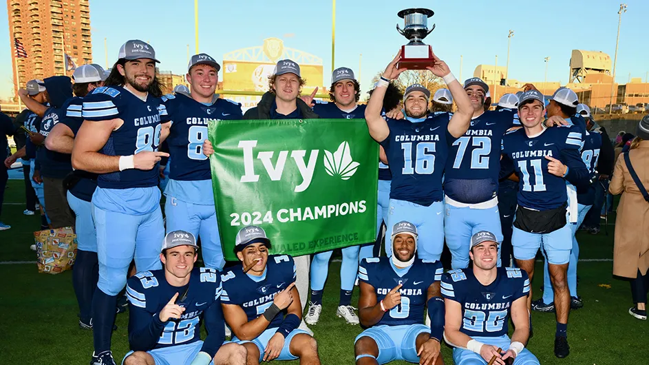 Several Columbia football players hoist an Ivy League championship trophy and hold a championship banner in celebration at Columbia's Baker Athletics Complex.