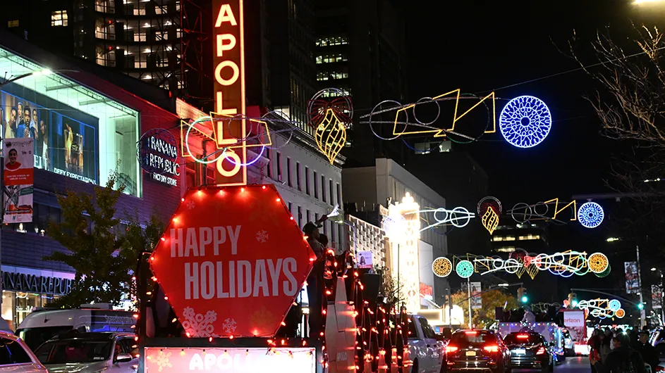 Harlem Holiday Parade floats with festive displays of Happy Holidays ride by the illuminated Apollo Theater marquee under big holiday-decorated street ornaments on 125th Street in New York City. 
